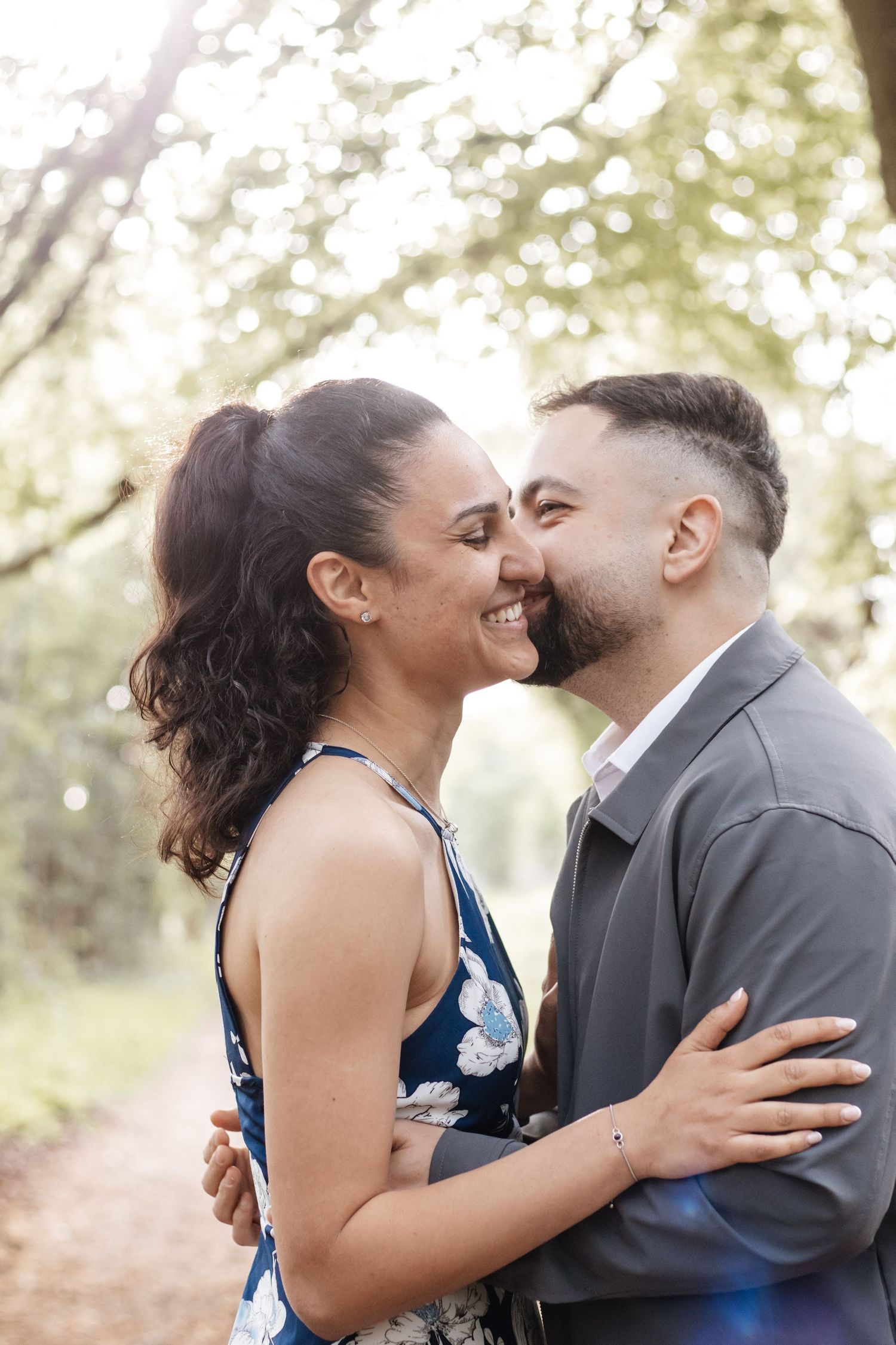 A couple shares a romantic kiss outdoors, surrounded by blooming trees in soft natural light.