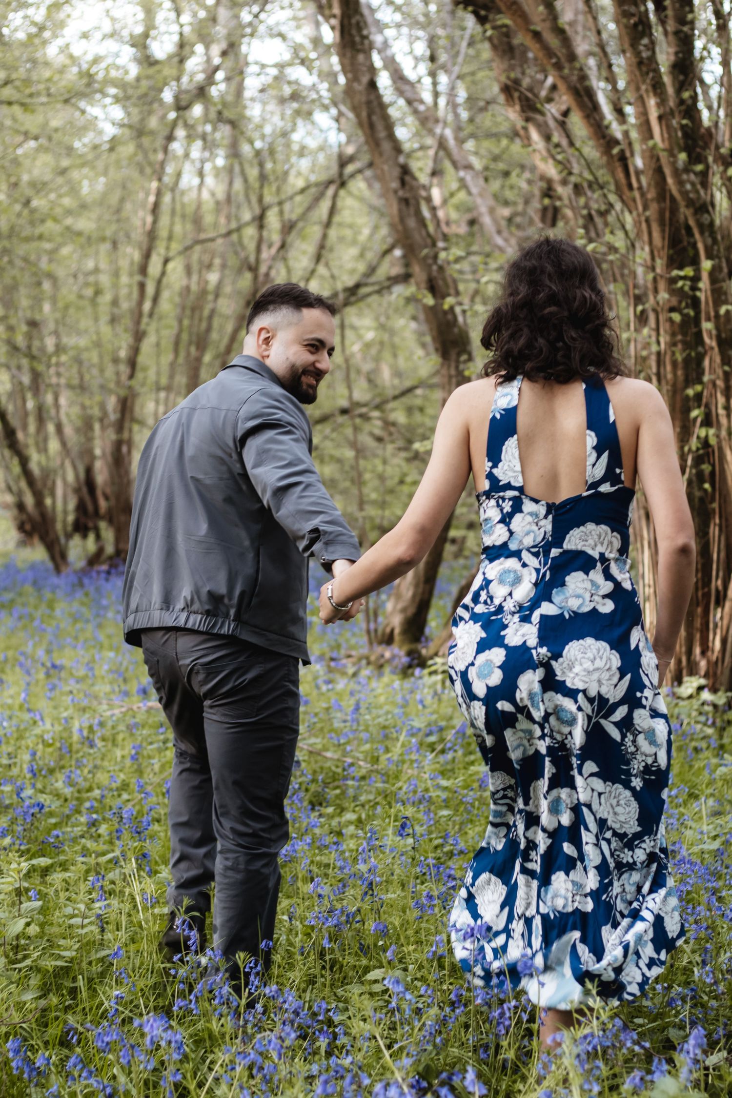 A couple walks hand-in-hand through a bluebell woodland, the woman wearing a floral navy dress and the man in a grey jacket.