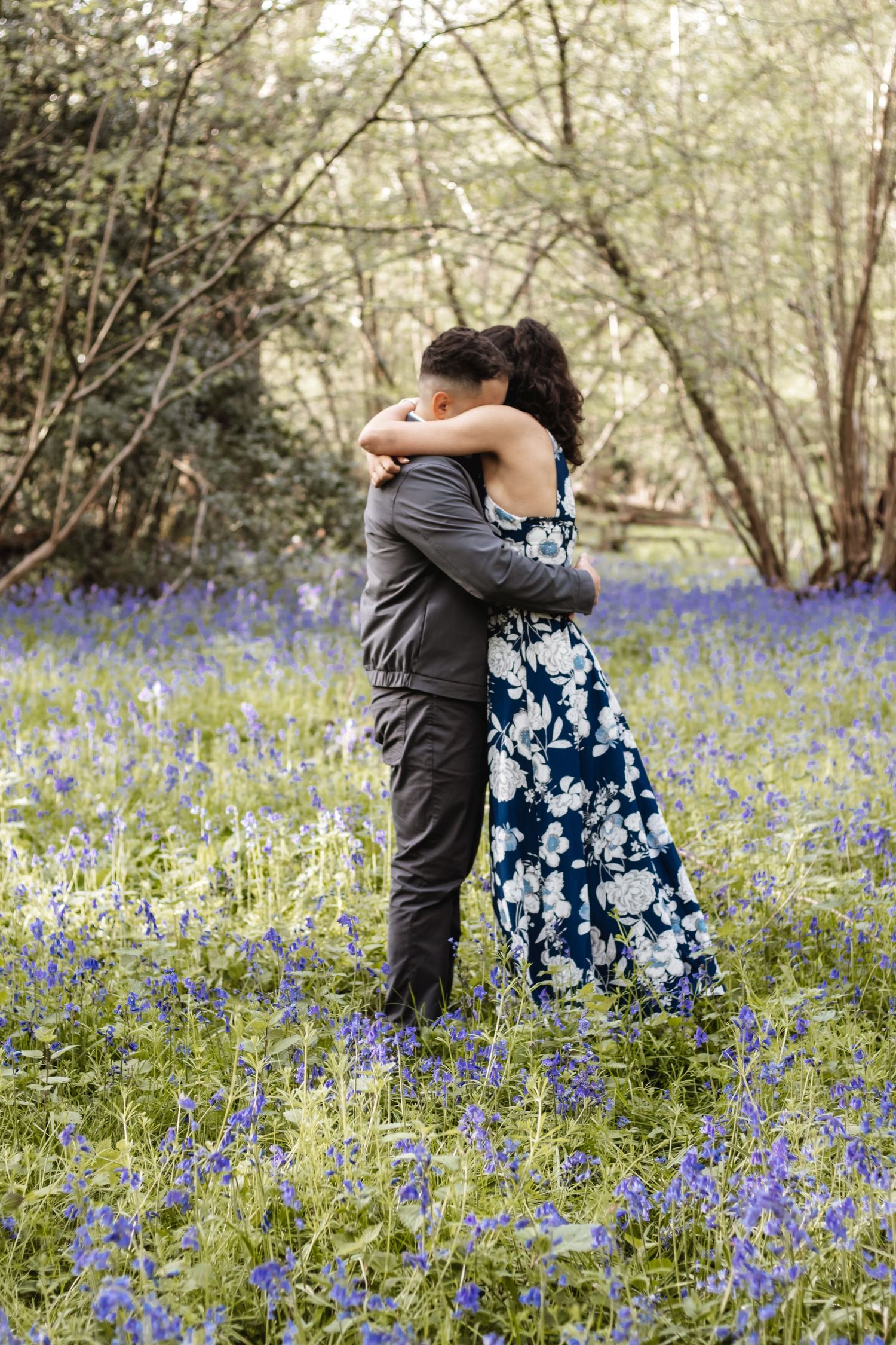 Couple embracing in a bluebell woodland, woman in floral blue dress, man in grey suit, surrounded by purple wildflowers.
