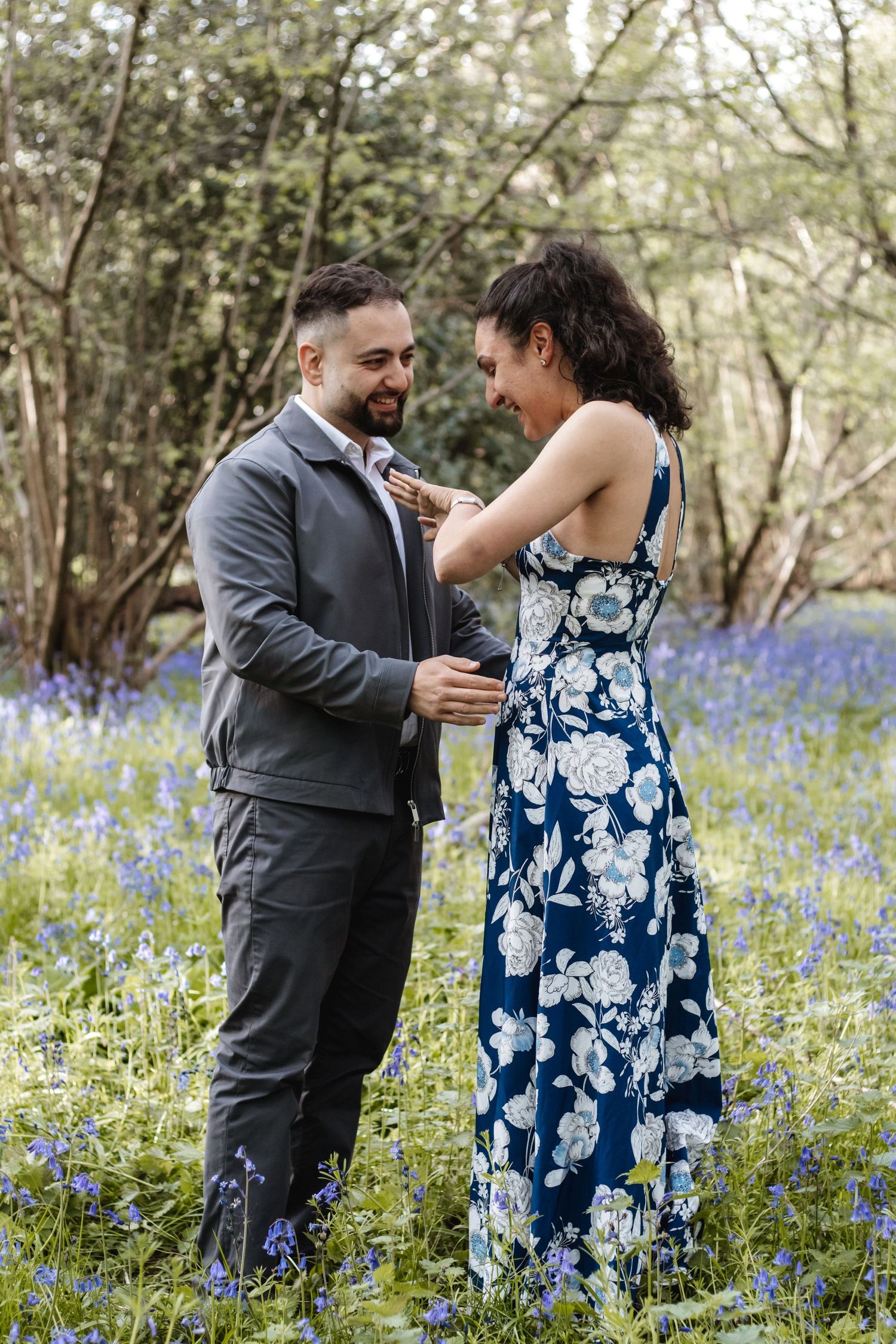 Couple smiling in a bluebell woodland, woman in floral dress adjusting mans suit jacket outdoors.