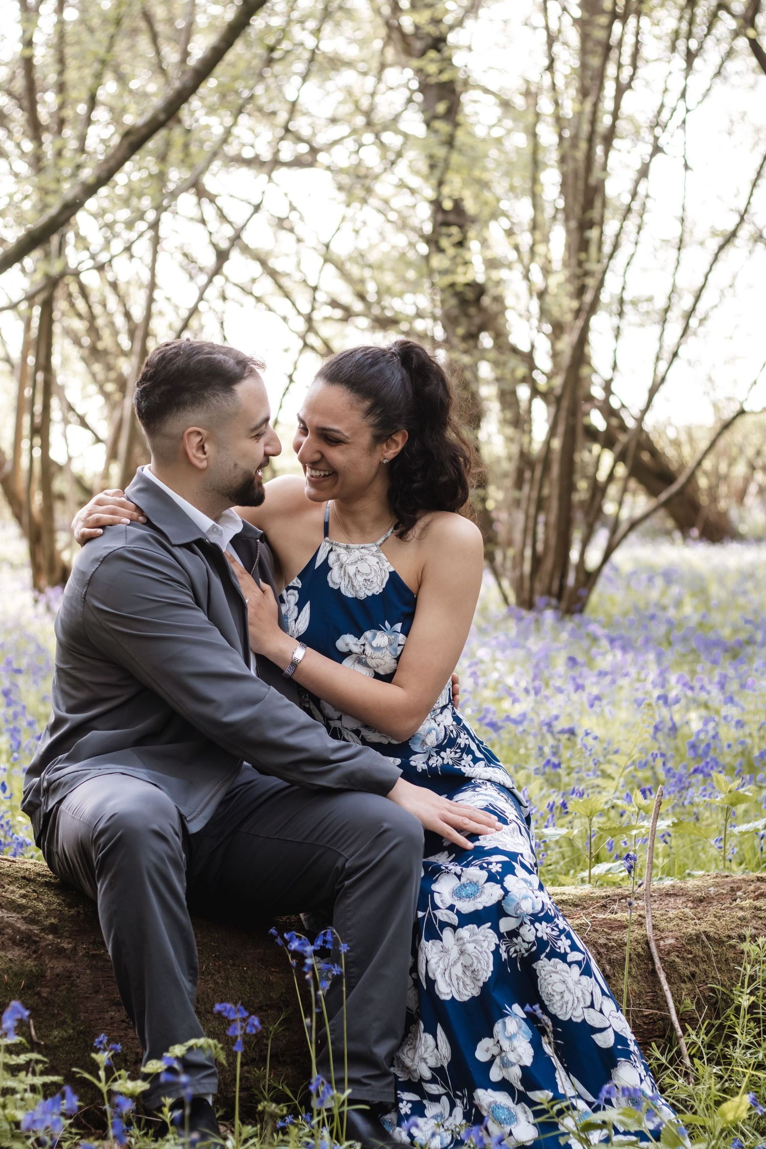 Couple sitting on a log in a bluebell woodland, smiling at each other, woman in floral navy dress, man in grey suit.