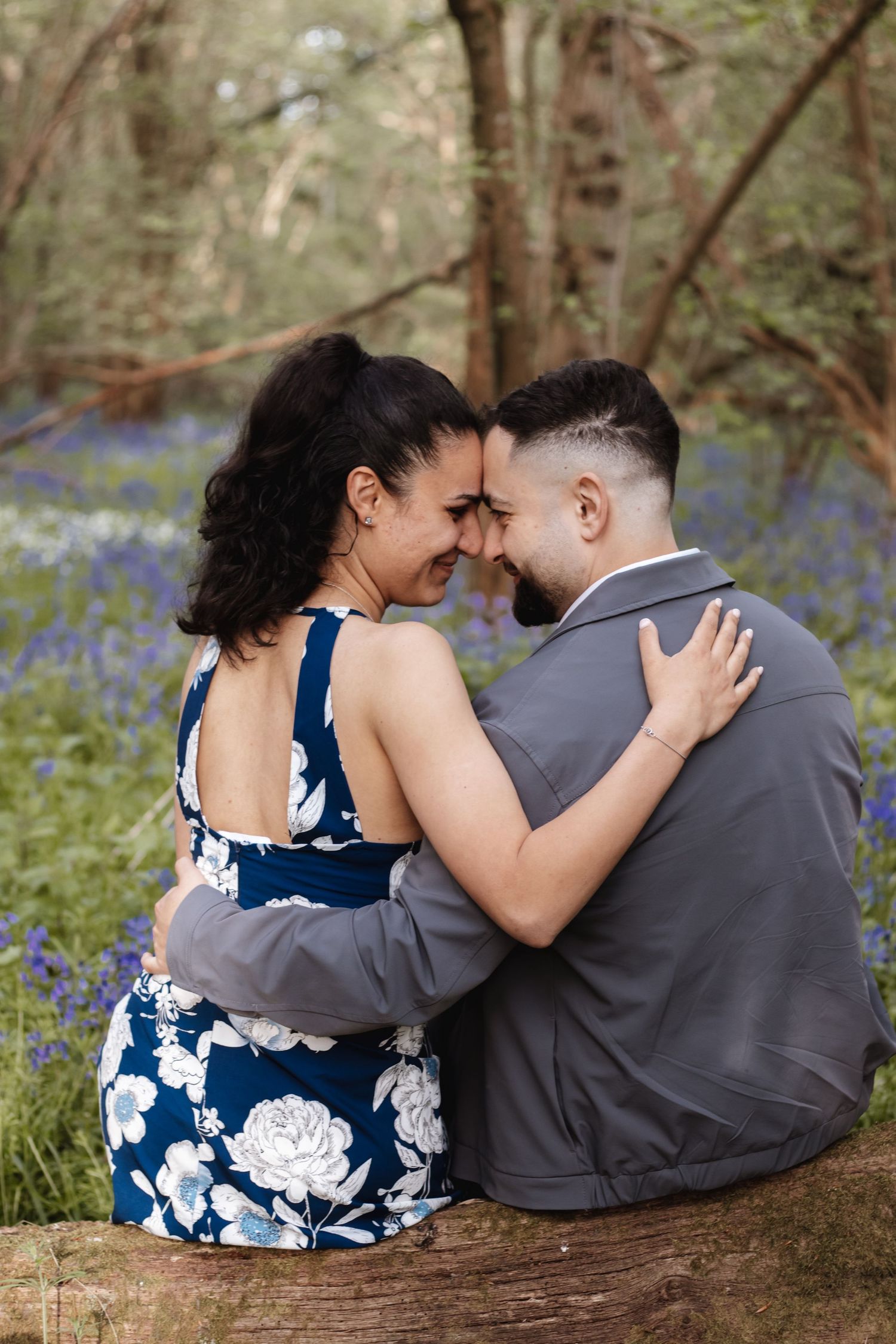 Couple sitting on a log in a bluebell woodland, touching foreheads and embracing romantically.
