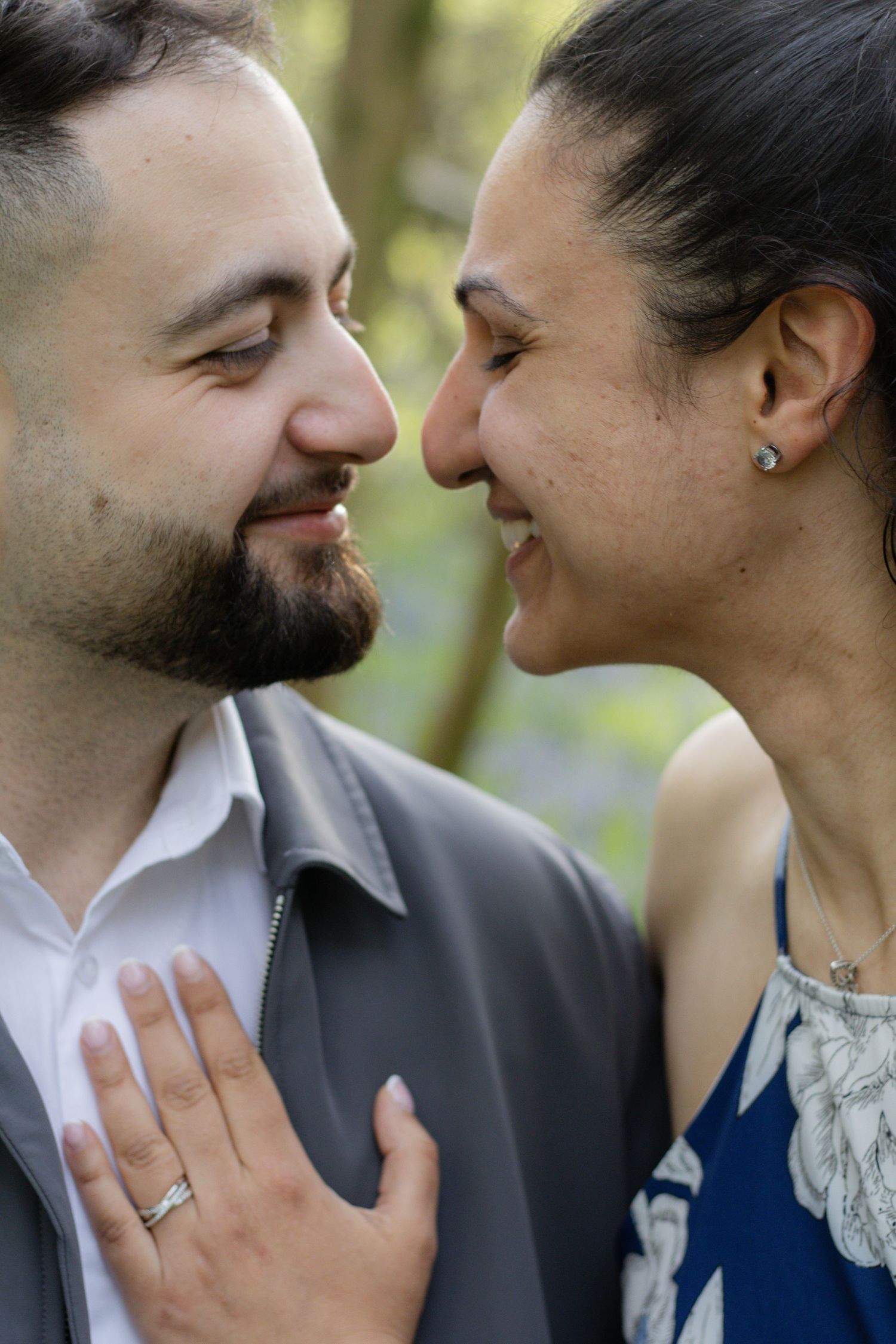 Couple sharing a romantic moment outdoors, woman smiling and placing hand on mans chest, wearing engagement ring.