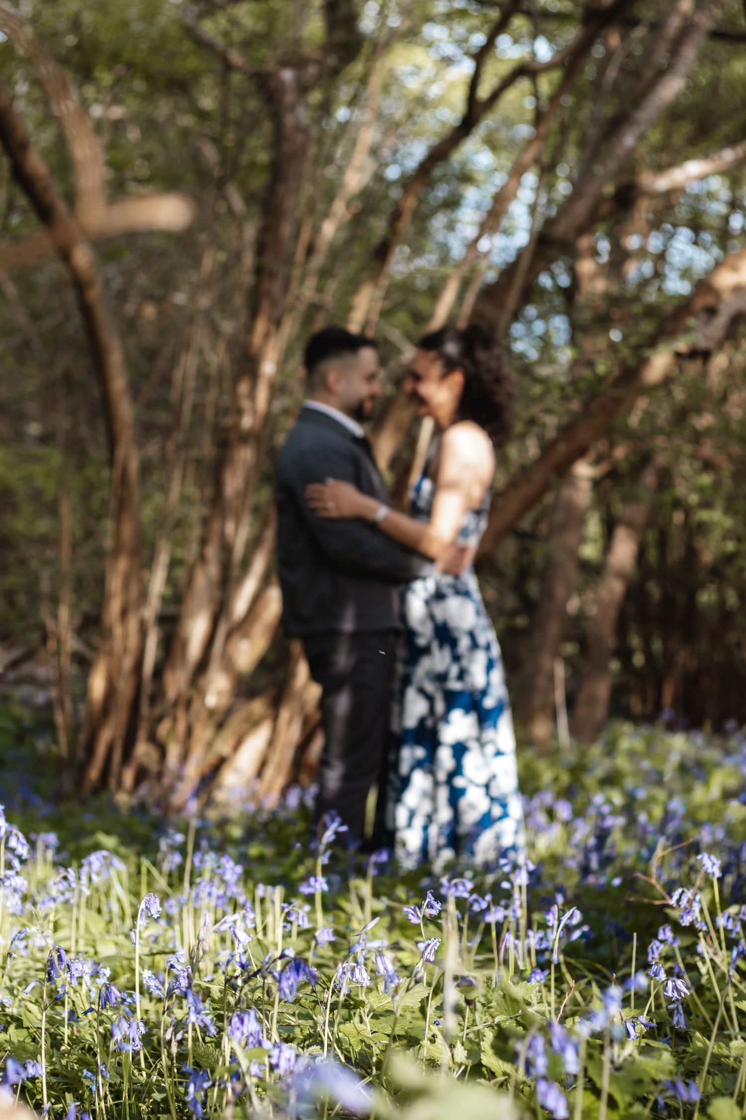 Couple embracing in a bluebell woodland, woman in floral dress, soft focus foreground of purple wildflowers.