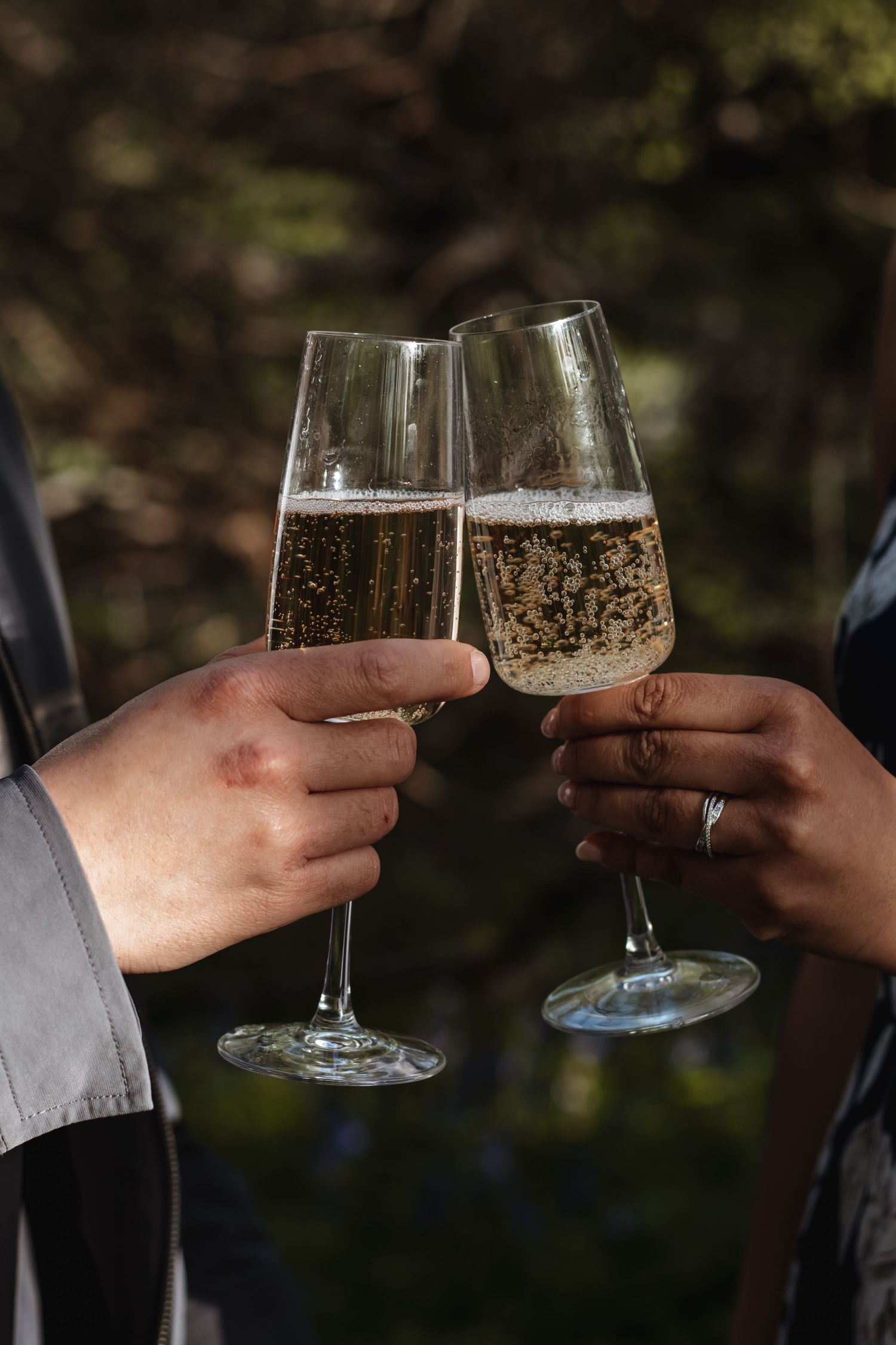Two people clink champagne flutes in a celebratory toast outdoors, with one hand wearing an engagement ring.