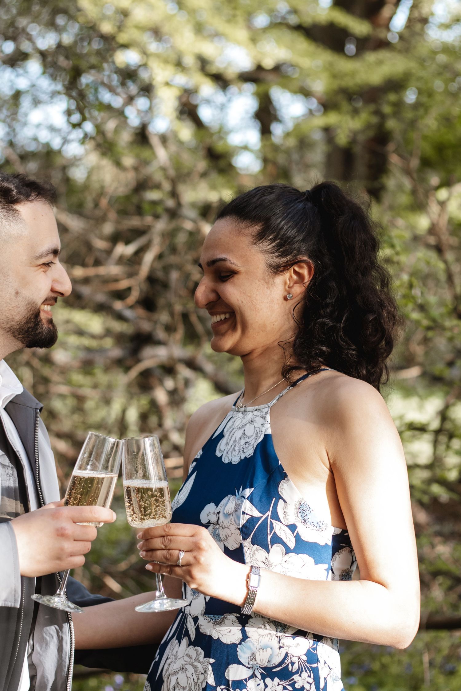 Couple toasting with champagne flutes outdoors, smiling joyfully, woman in blue floral dress, trees in background.