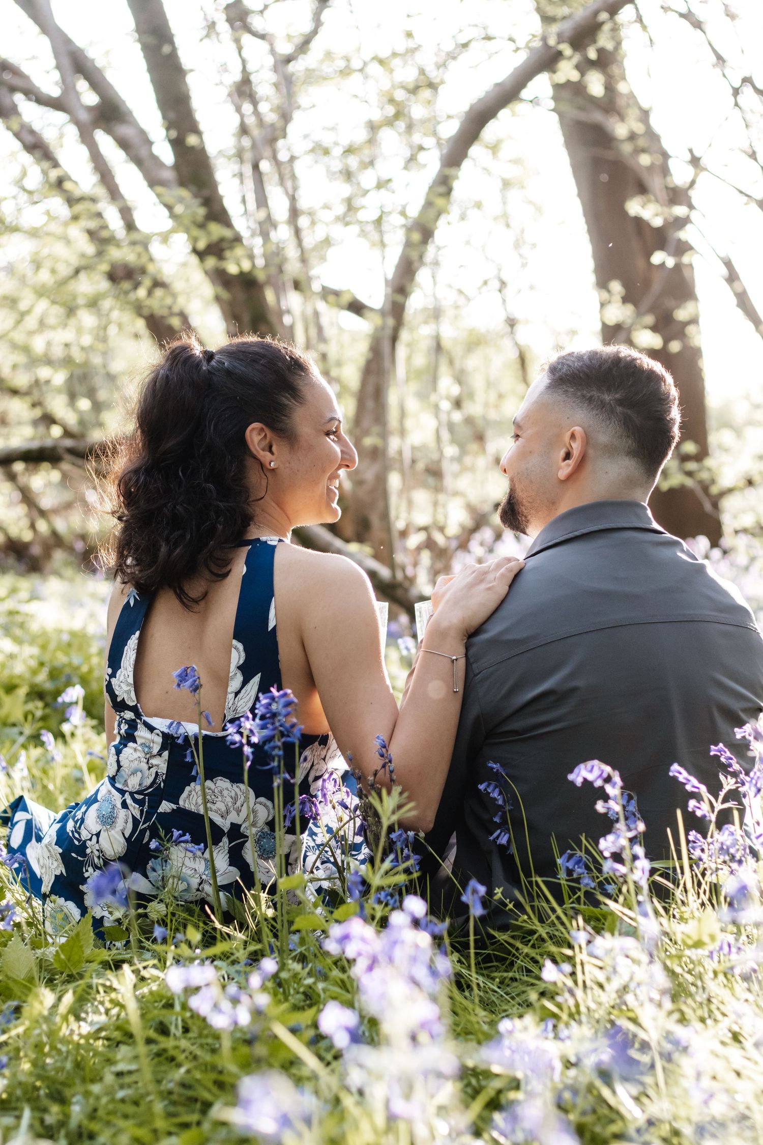 Couple sitting in a field of purple wildflowers, smiling at each other, surrounded by sunlit trees in bloom.