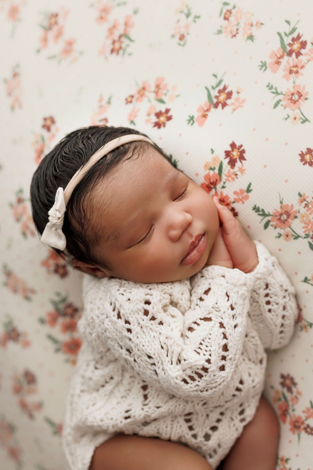 Sleeping newborn baby girl in white knit outfit and bow headband on floral background.