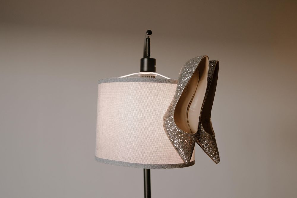 Glittery silver high heel shoes resting on a white lamp shade on a floor lamp stand.
