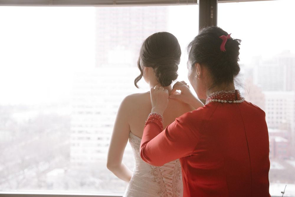 A mother in red helps her daughter in a white wedding dress with a necklace by a bright window overlooking a city.