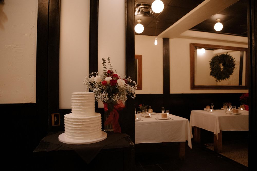 Elegant white tiered wedding cake beside floral arrangement in candlelit restaurant dining room with wreath decor.