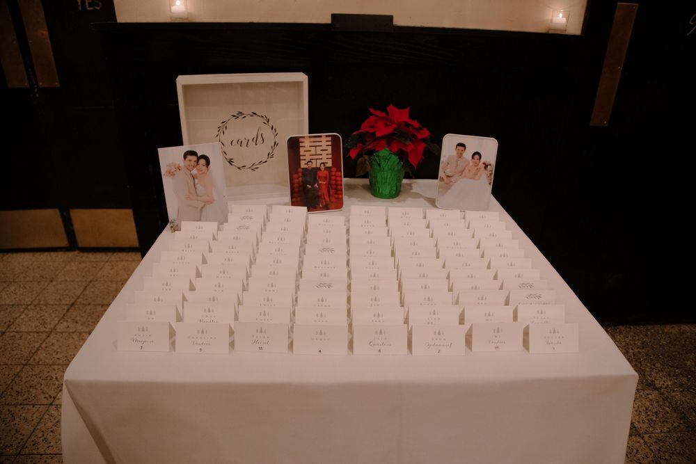 Wedding escort card table with white place cards, floral wreath sign, red flower centerpiece, and photo displays at reception.