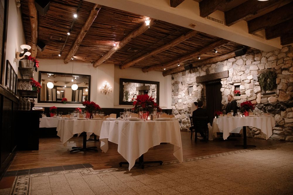 Elegant restaurant dining room with white tablecloths, stone walls, wooden beam ceiling, and red poinsettia centerpieces.