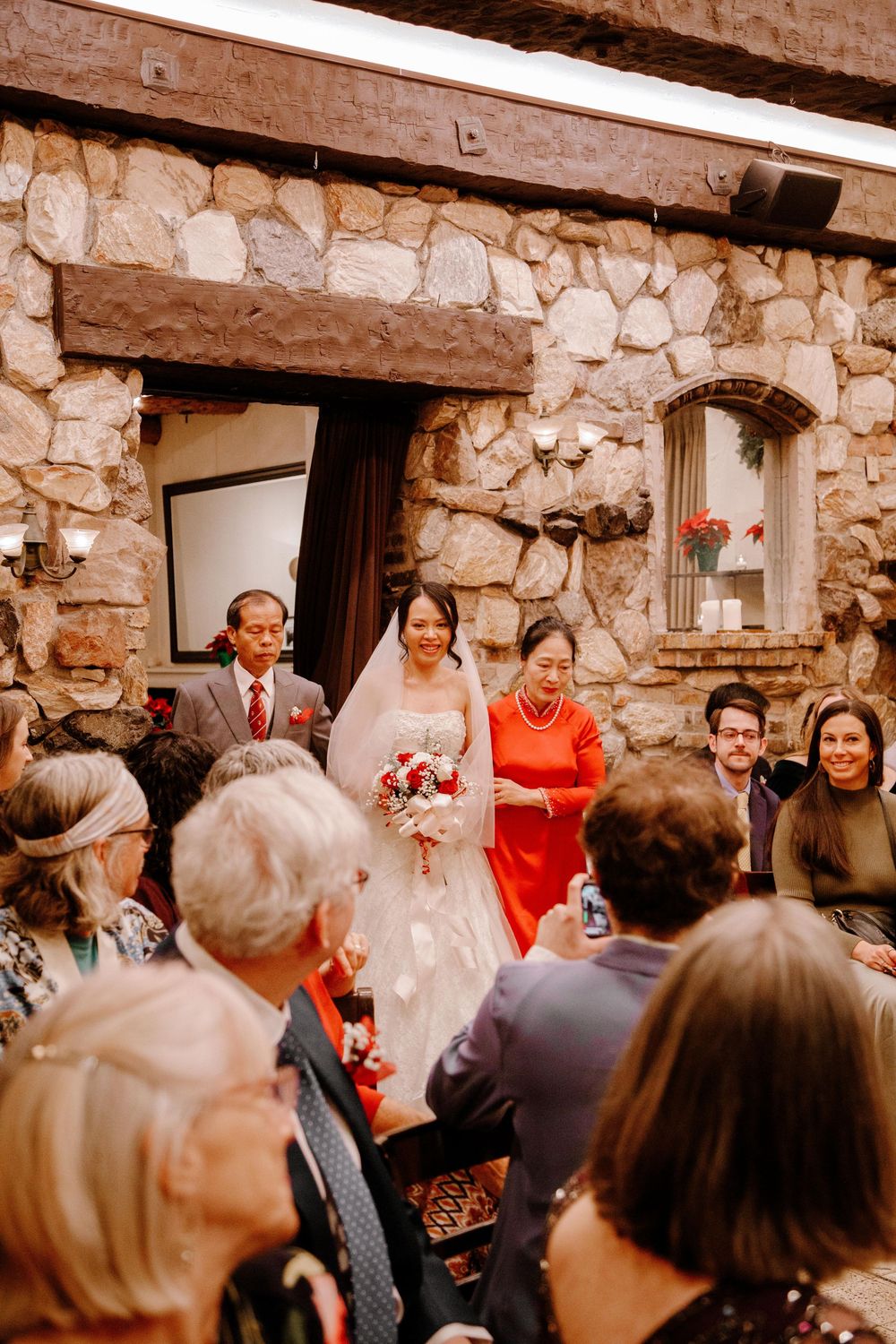 Bride walks down the aisle with family in a stone-walled wedding venue, wearing a white gown and holding a red bouquet.