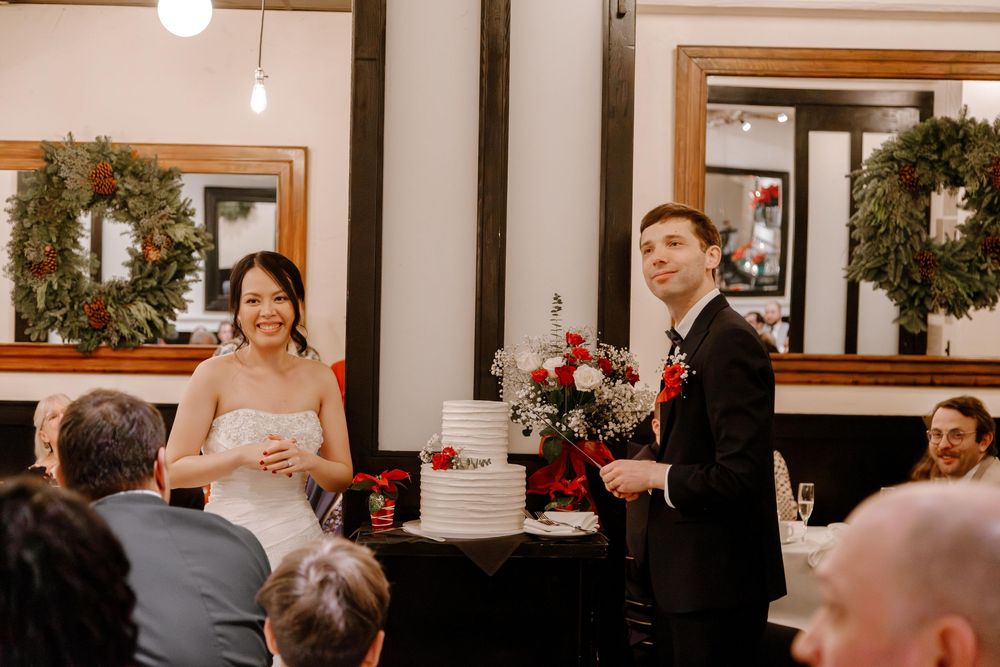 Bride and groom standing by white wedding cake with red floral decor, wreaths on mirrors in background.