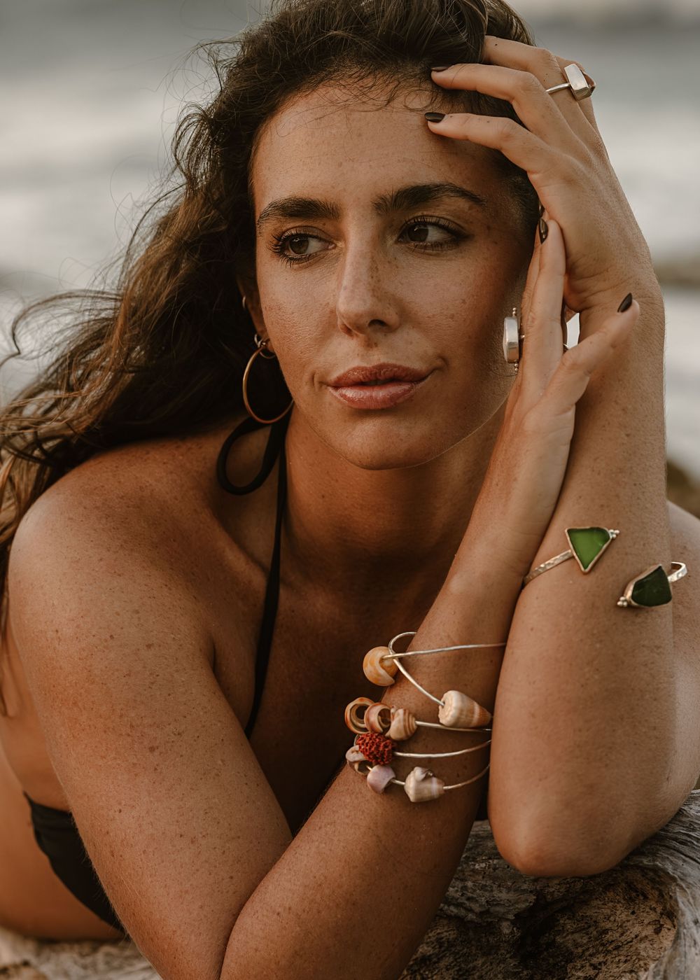 Woman in black bikini poses on beach wearing layered bracelets and rings, with wavy brunette hair and freckles.