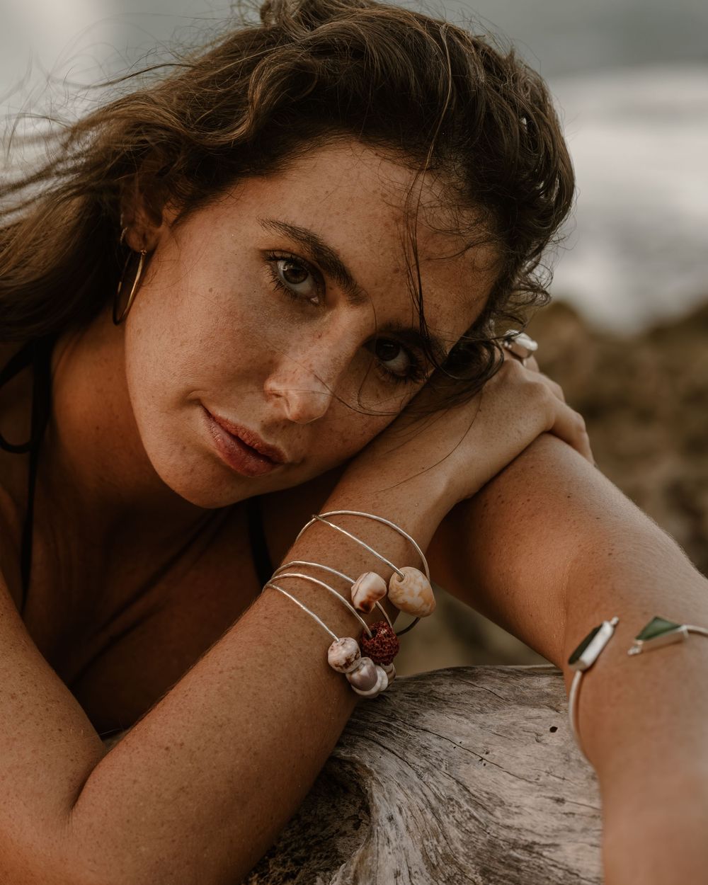 Woman with freckles wearing shell bracelets poses on rocky beach, her chin resting on hands.