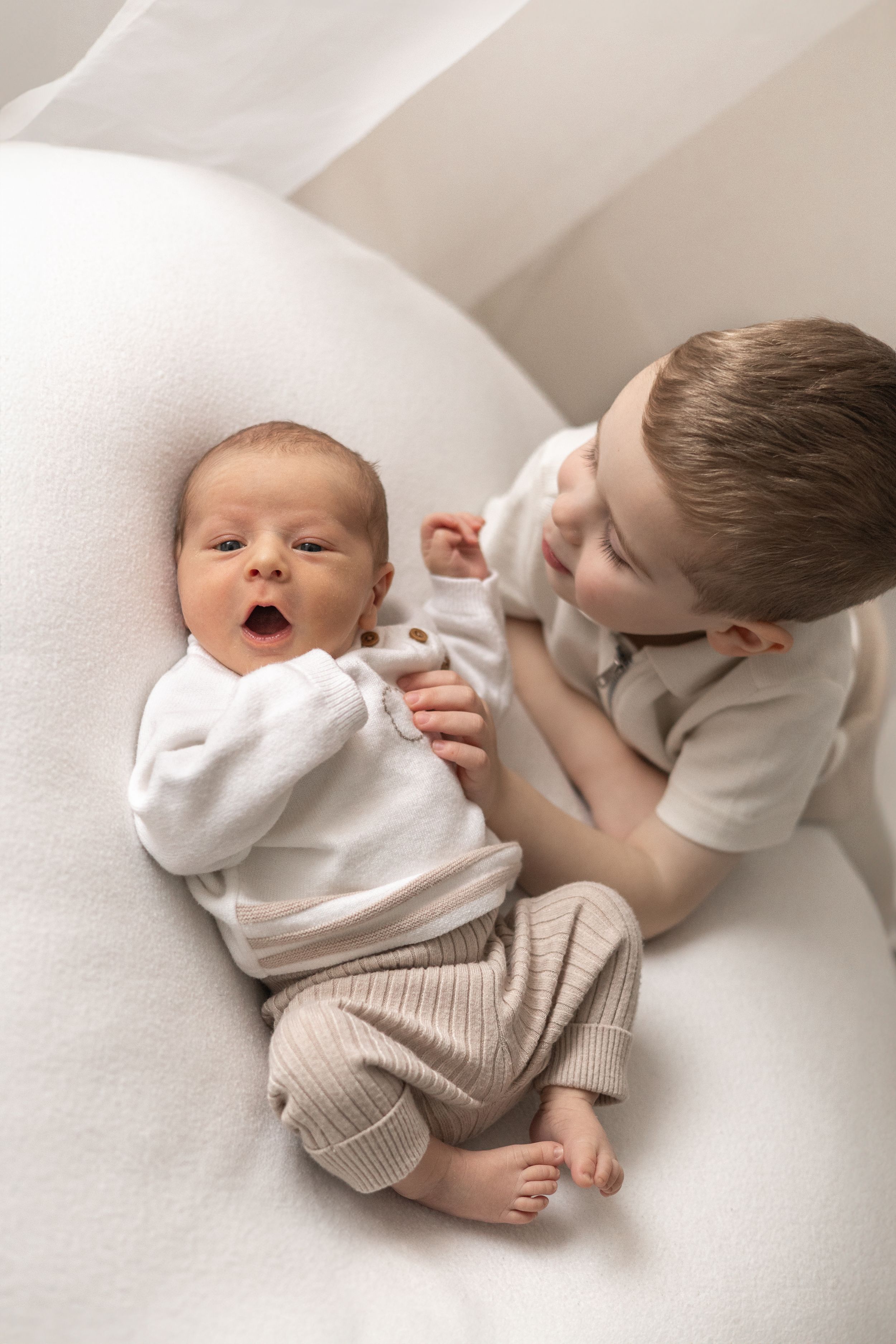 Older sibling gently interacting with newborn baby sibling lying on white cushion baby photographer Nottingham newborn session