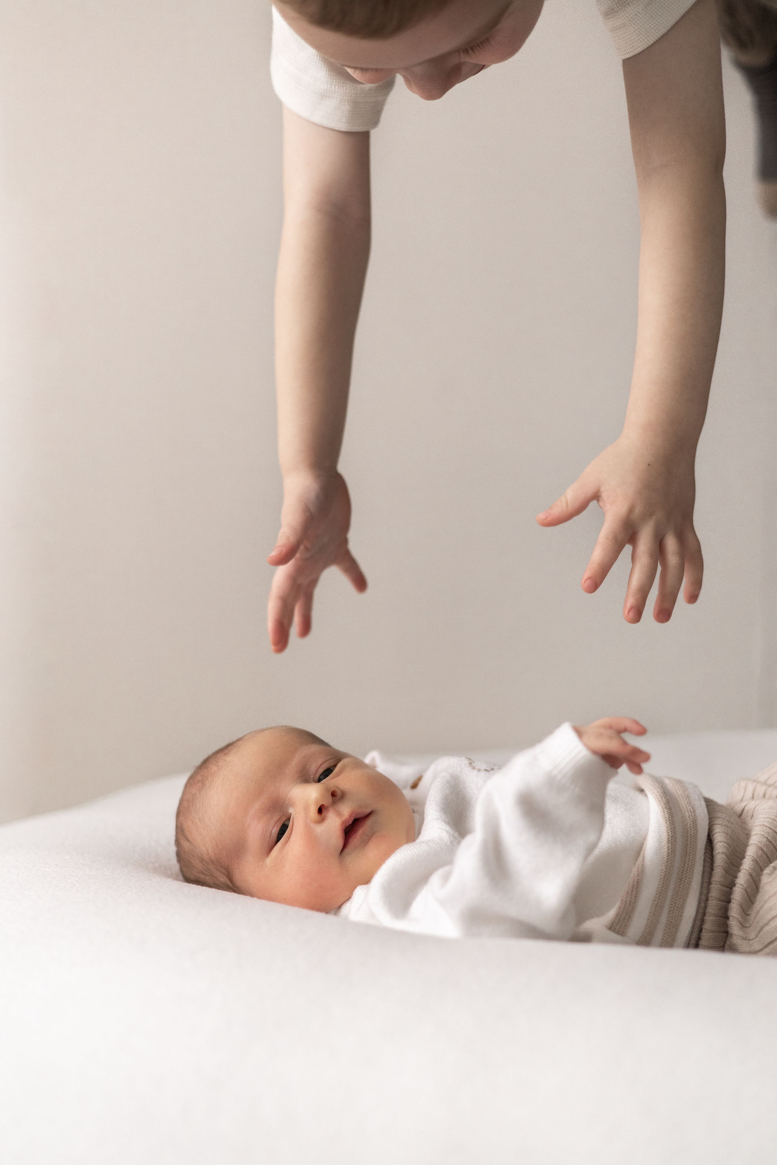 Toddler reaching down toward newborn baby lying on white surface from above baby photographer Nottingham newborn session