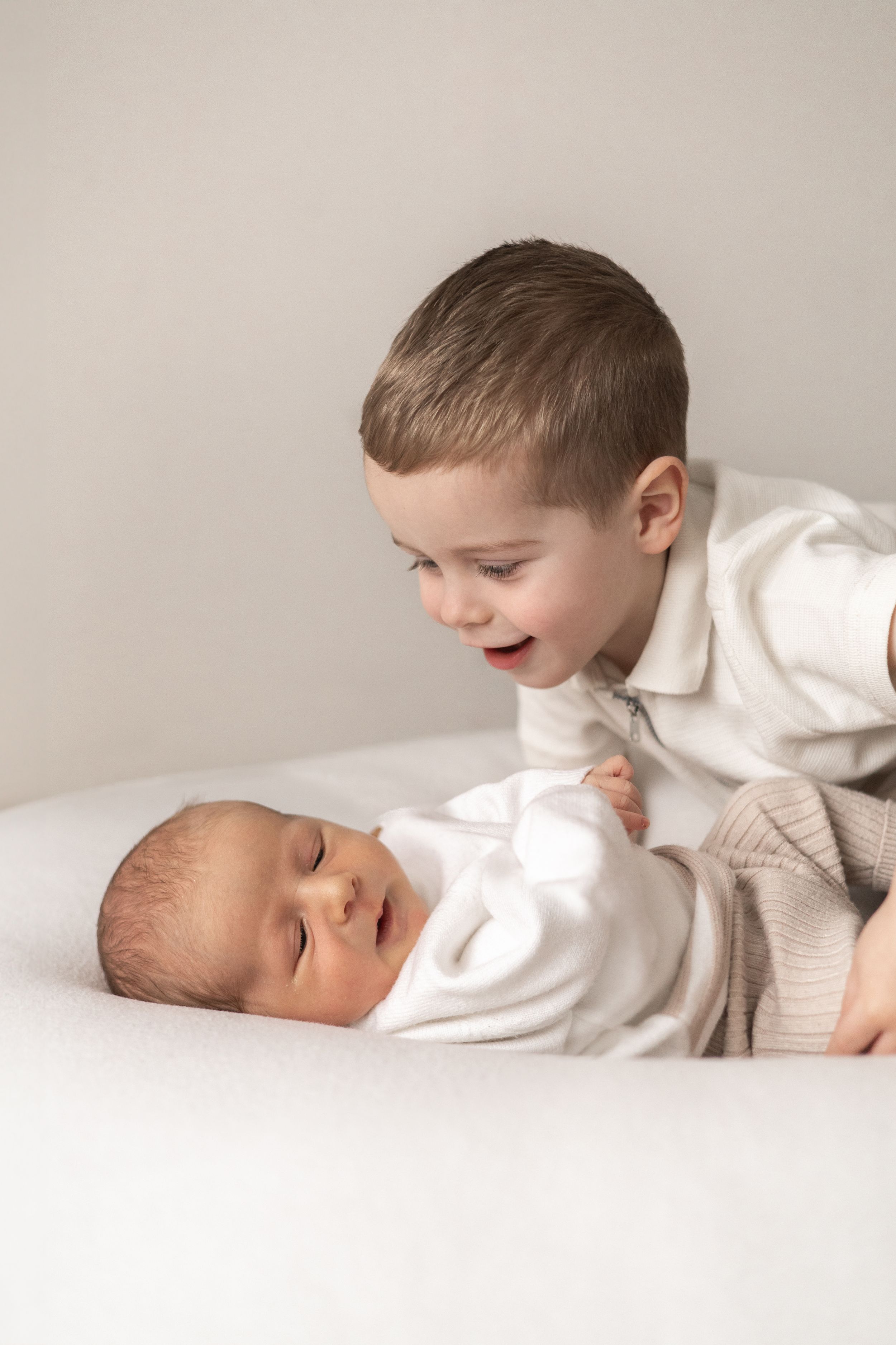 Excited young sibling leaning over a newborn baby on a white surface, both dressed in neutral tones baby photographer Nottingham newborn ses