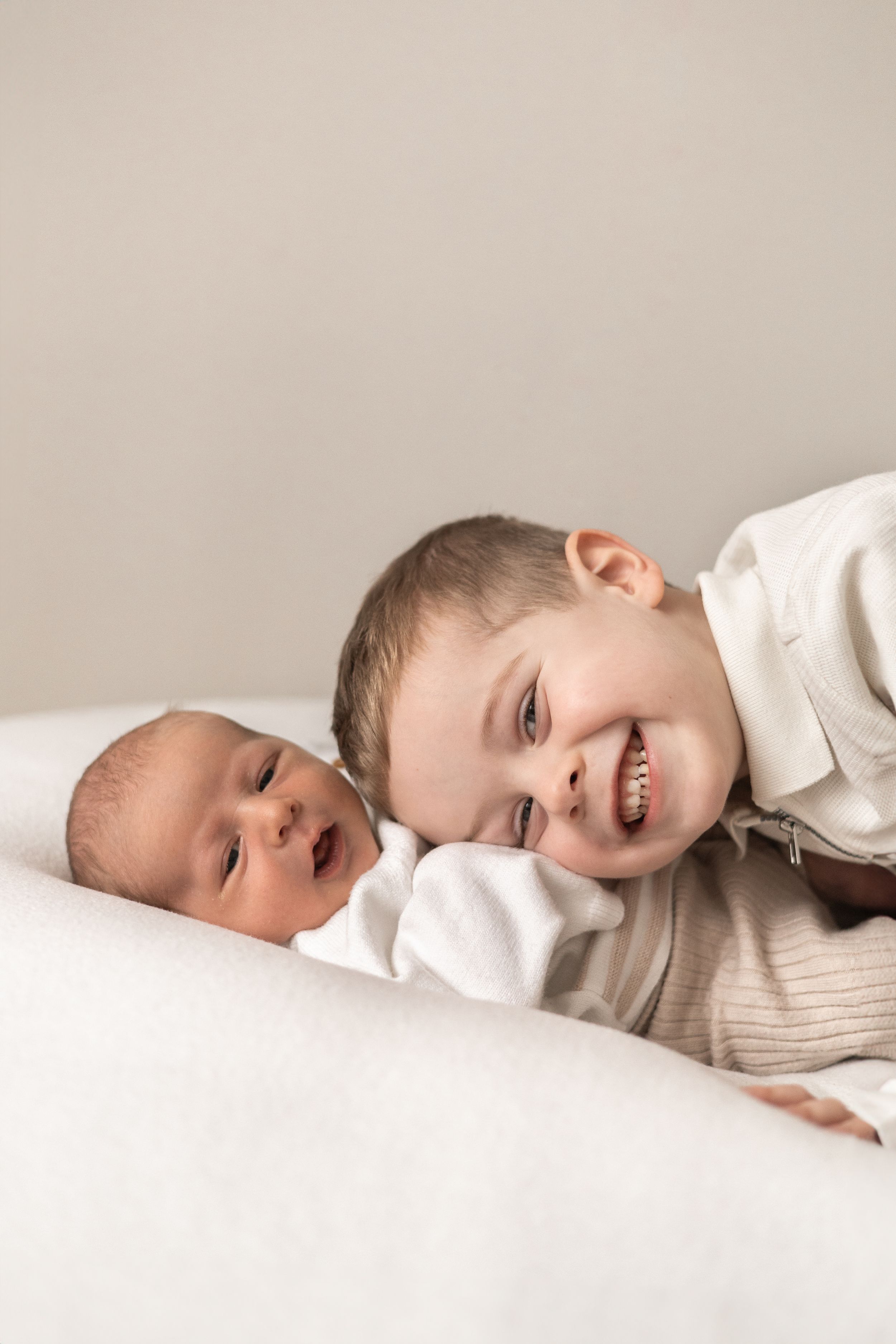 A smiling young sibling lying next to a newborn baby on a white surface, sibling portrait photo baby photographer Nottingham newborn session