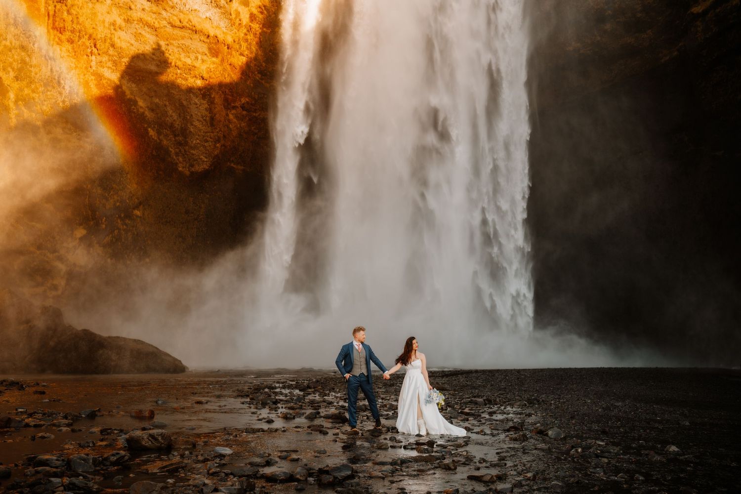 Bride and groom hold hands before a massive waterfall with golden light and a rainbow in Iceland.