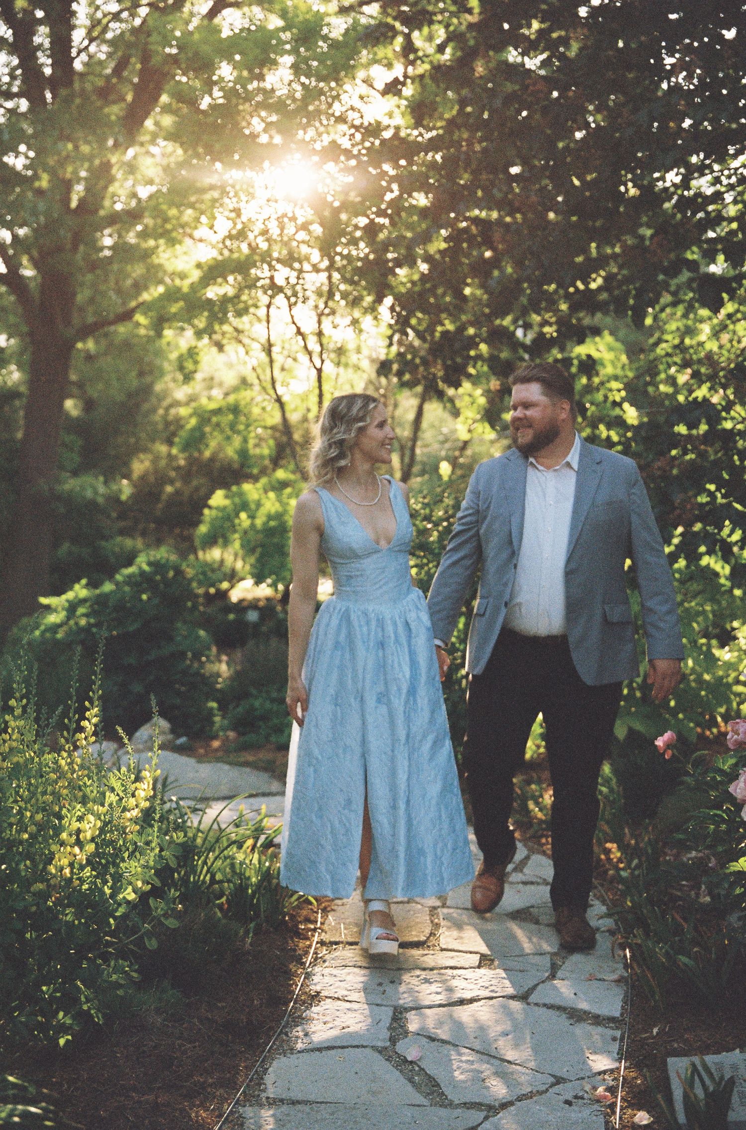 Couple smiling at each other in a lush garden greenhouse with colorful pink flowers and green trees.
