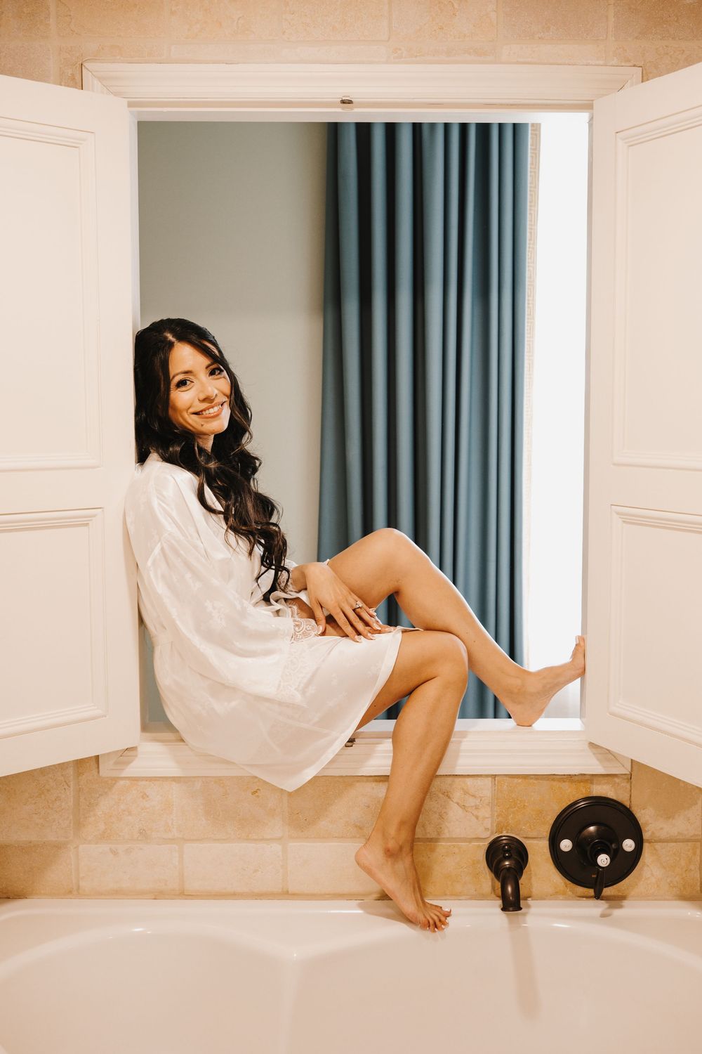 A smiling brunette woman in a white robe sits on a bathtub ledge near a window with blue curtains.