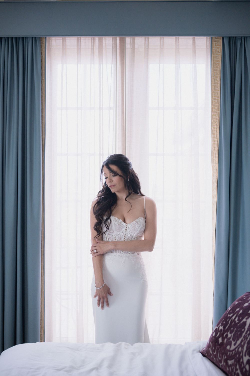 Bride in white lace wedding gown standing by a sunlit window in a hotel room, looking down elegantly.
