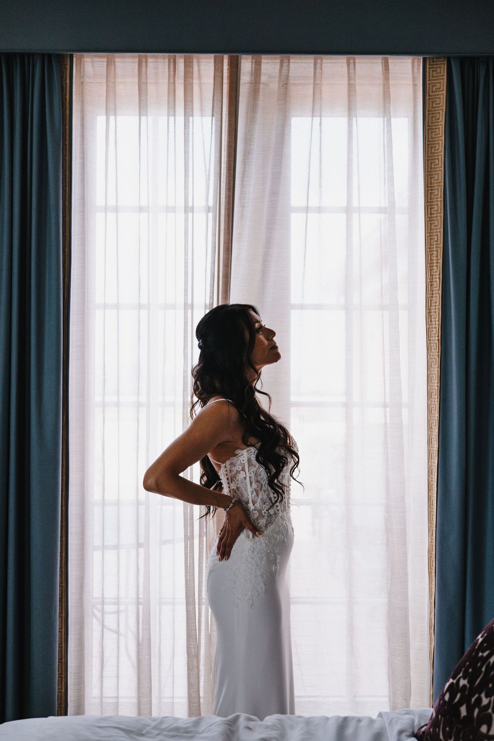 Bride in lace wedding gown poses by sheer curtained window in soft backlit bridal suite.