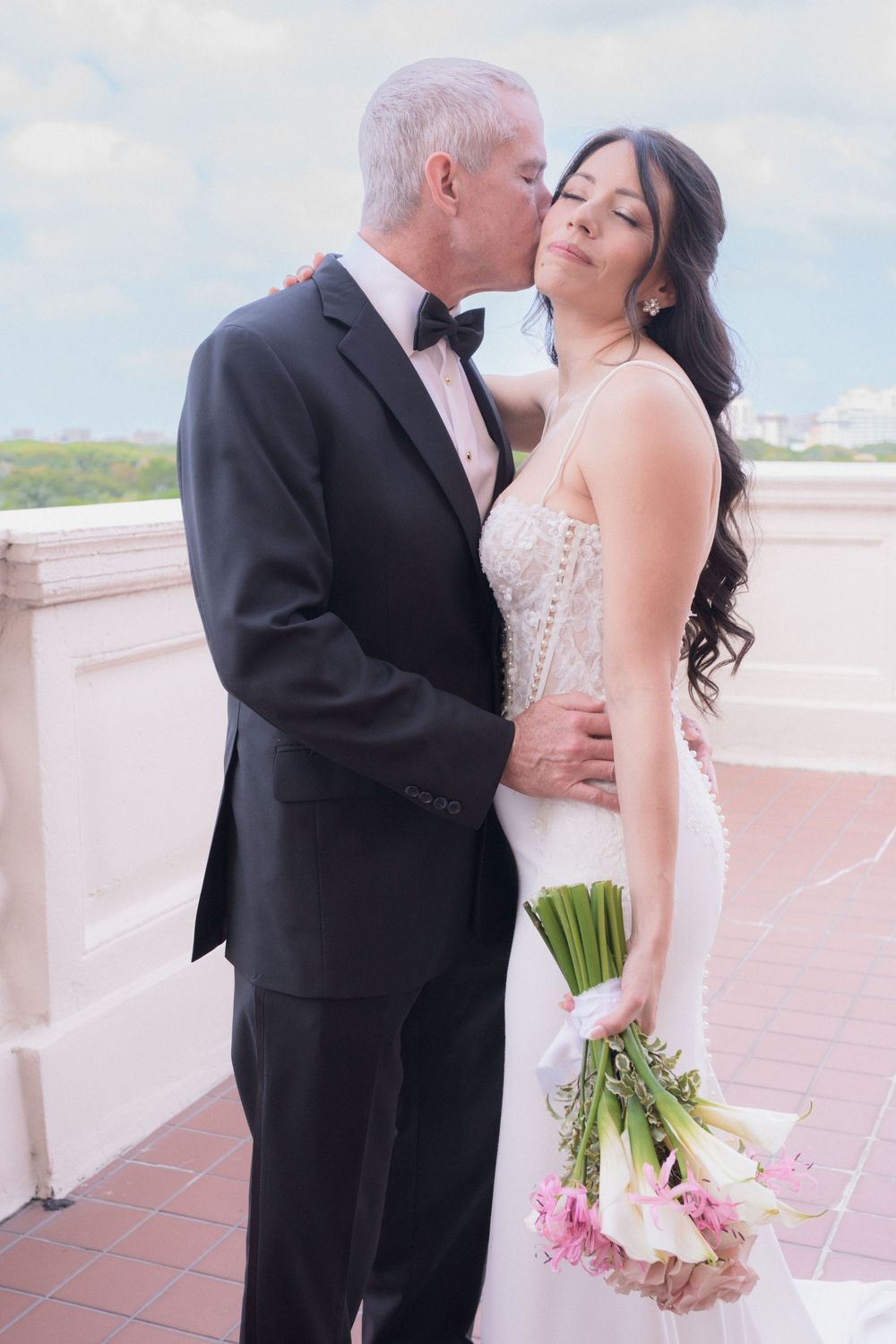 A groom in a black tuxedo kisses his bride in a white gown holding a pink and white bouquet on an outdoor terrace.