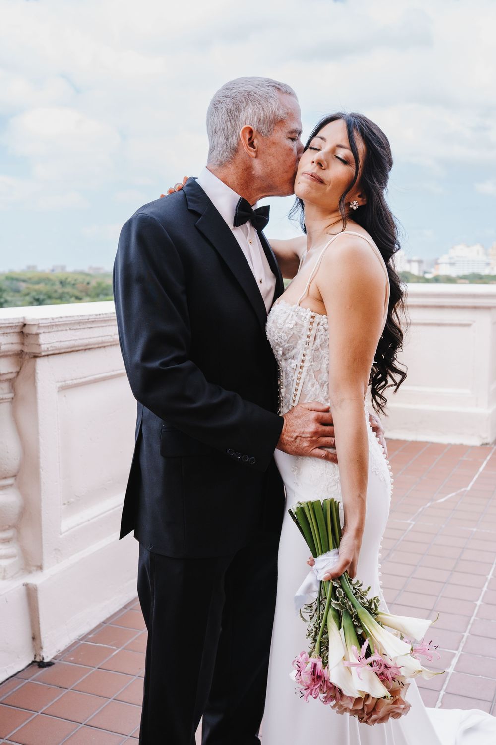 Bride and groom share a romantic kiss on an outdoor terrace, bride holding a white and pink bouquet in a strapless gown.