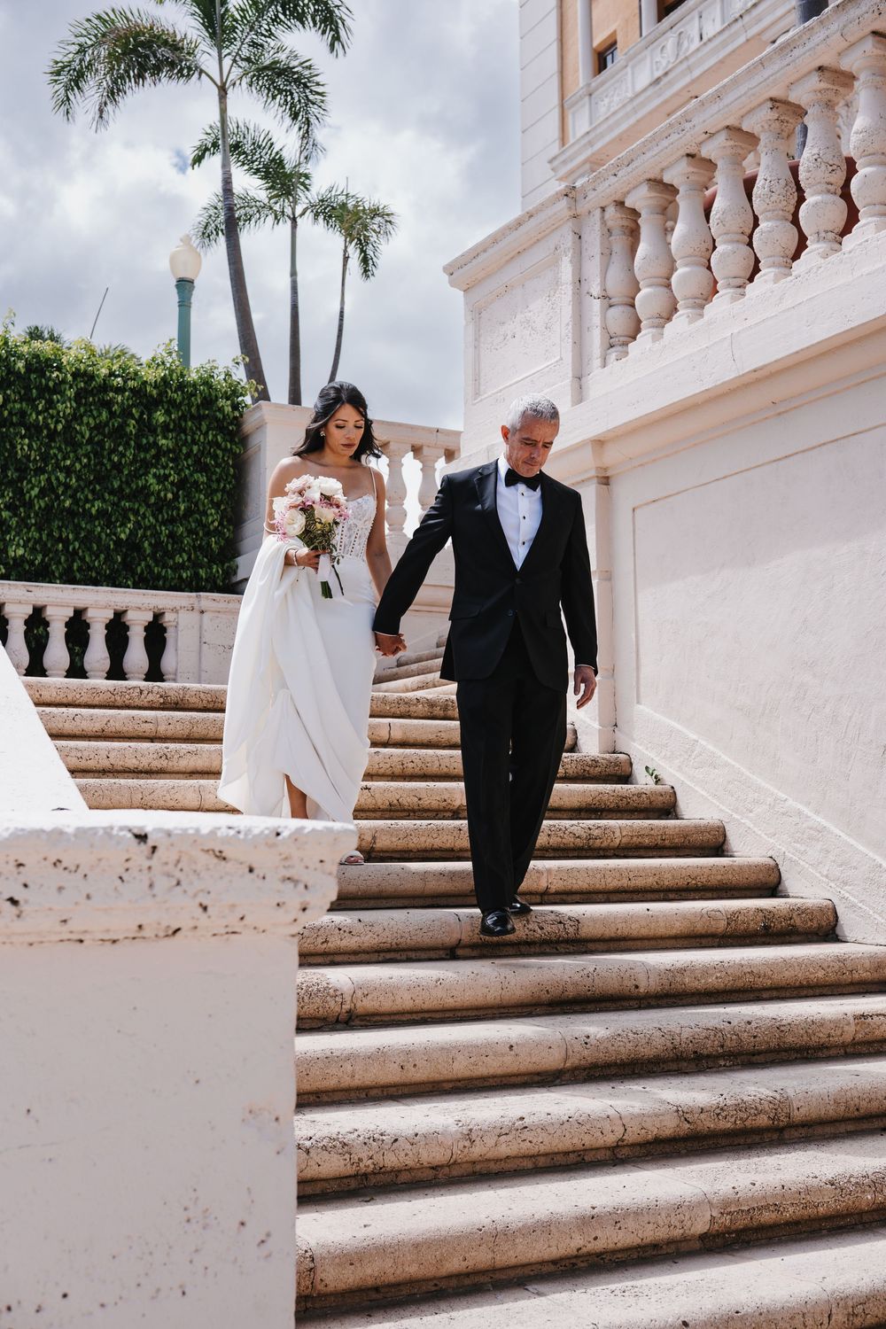 Bride and groom descend elegant stone staircase at outdoor wedding venue lined with palm trees.