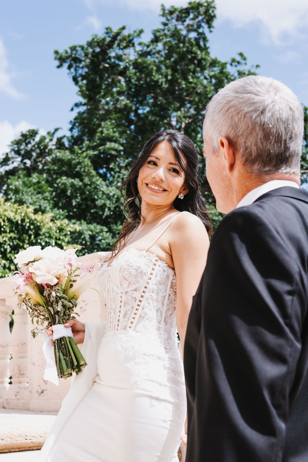 Smiling bride in white lace gown holds bouquet, facing older man in suit at outdoor wedding ceremony.
