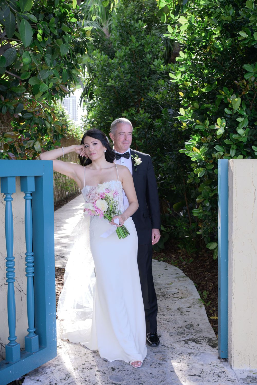 Bride in white gown holding pink bouquet stands with groom in tuxedo at blue garden gate.