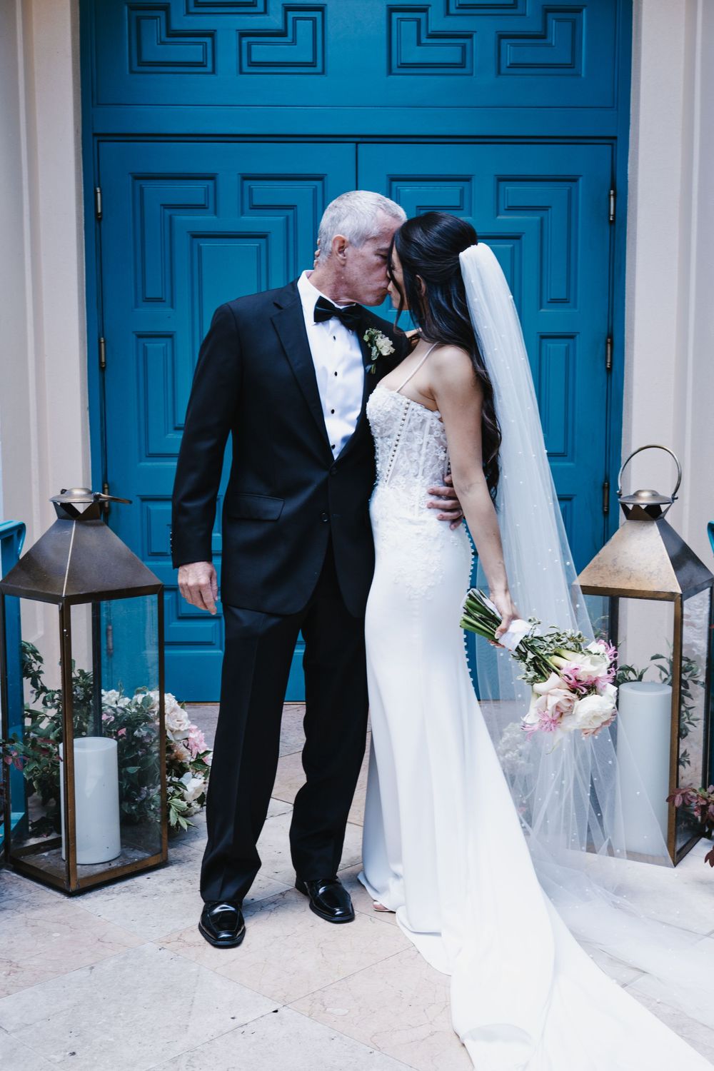Bride and groom share a kiss in front of a blue door, bride in white gown holding bouquet, groom in black tuxedo.