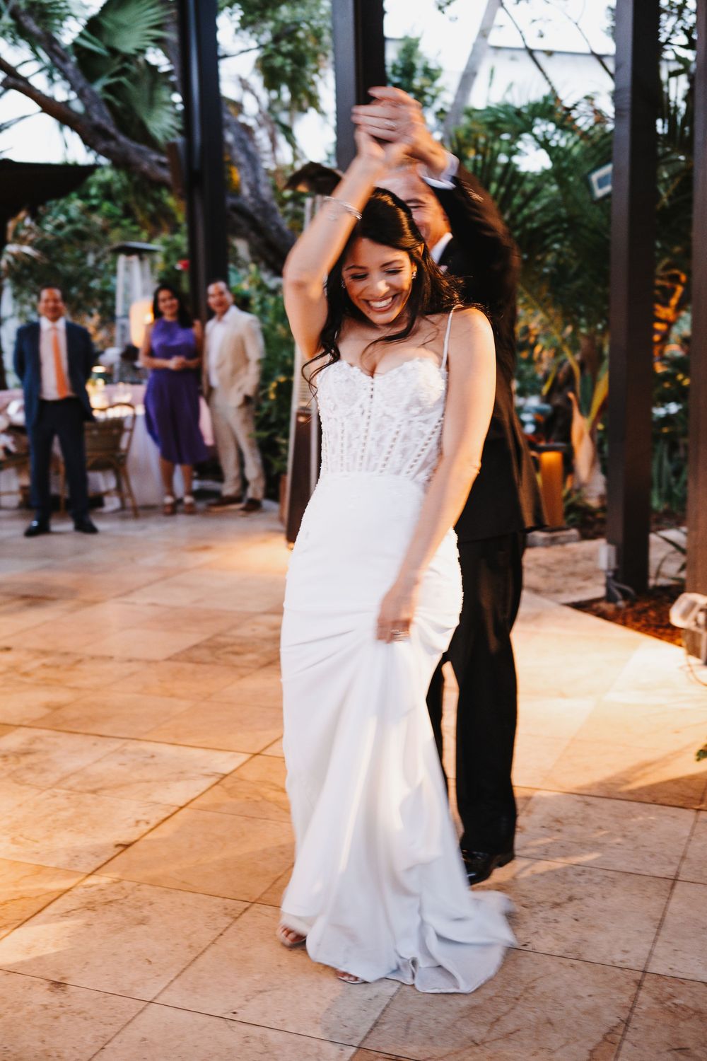 Bride in white gown laughs joyfully during first dance with groom at elegant outdoor wedding reception.