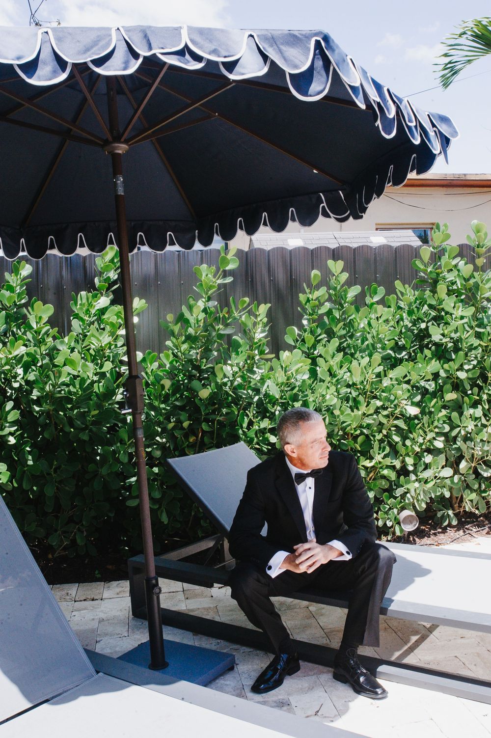 Groom in black tuxedo sits under a navy scalloped patio umbrella surrounded by lush green hedges outdoors.