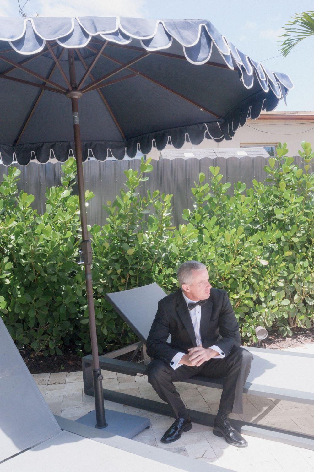 Man in tuxedo with bow tie sitting on bench under large blue patio umbrella surrounded by green hedges outdoors.