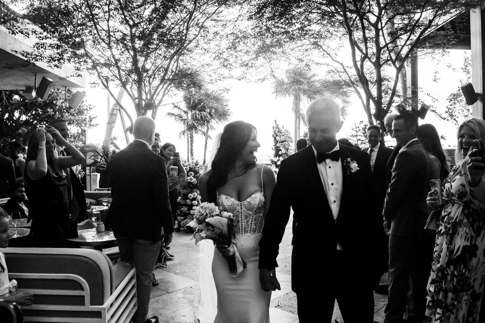 Black and white wedding photo of bride in strapless gown walking with groom in tuxedo through outdoor venue.