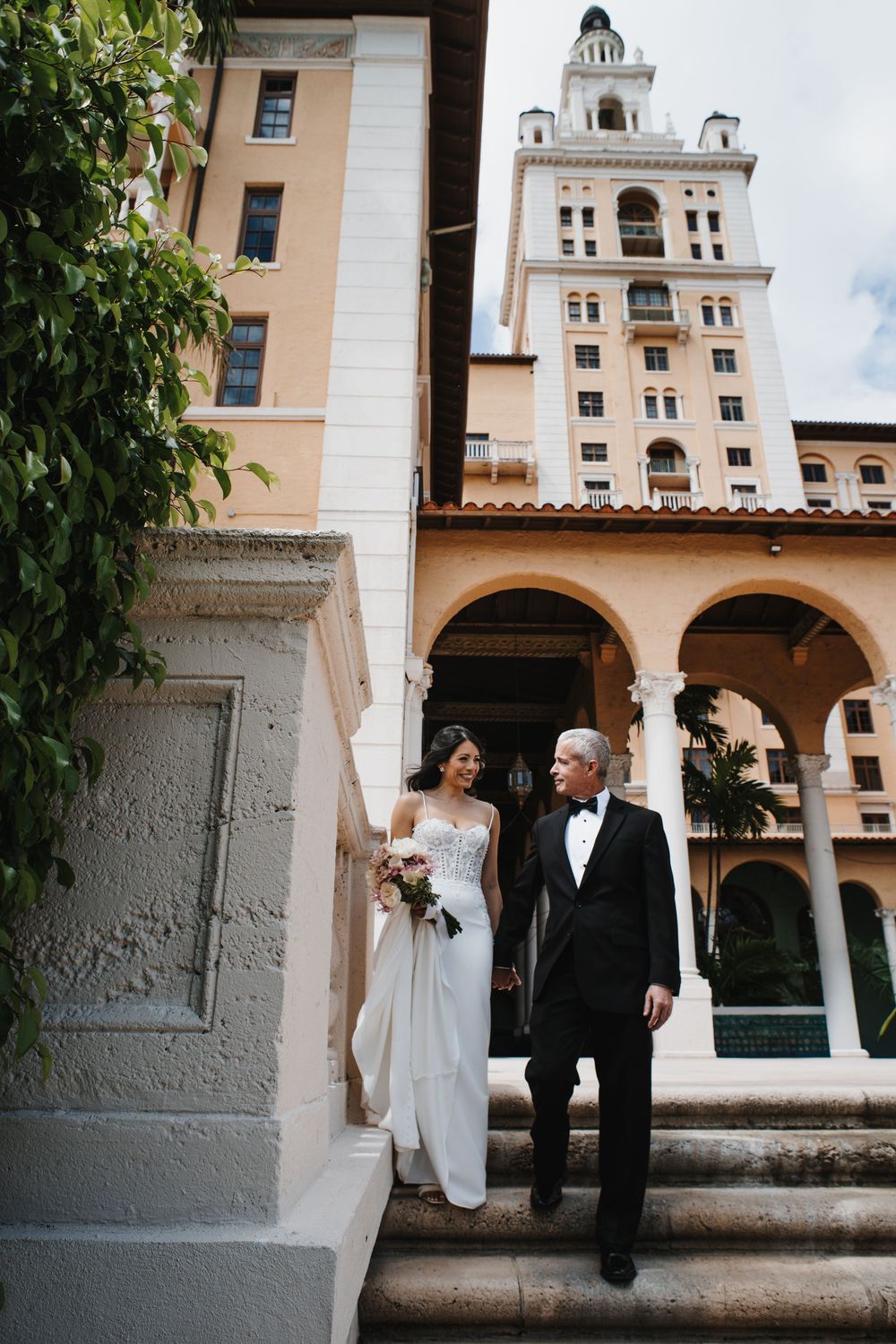 Bride in white gown and groom in tuxedo descend stone steps outside elegant hotel with tower.