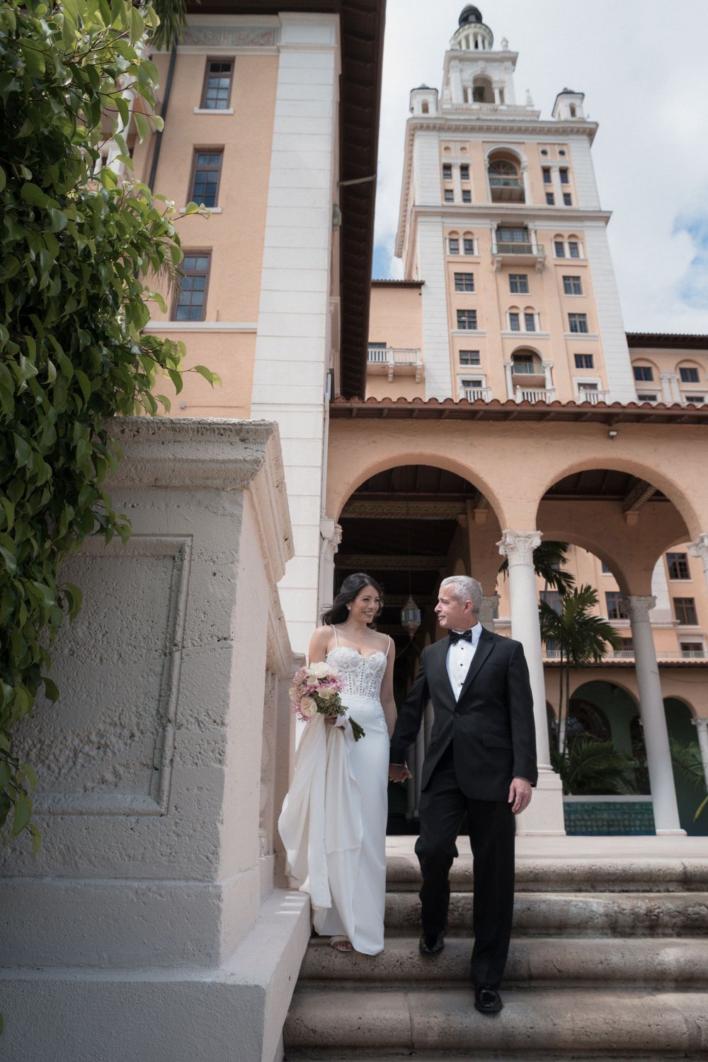Bride and groom in formal attire descend steps outside elegant hotel with tall tower.