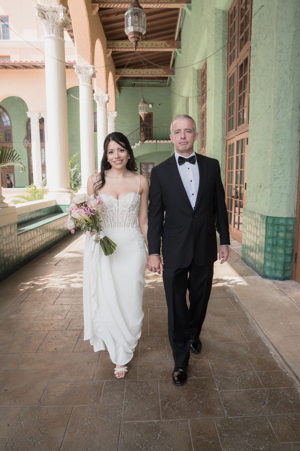 Bride in white gown and groom in black tuxedo walk hand in hand through elegant venue corridor.