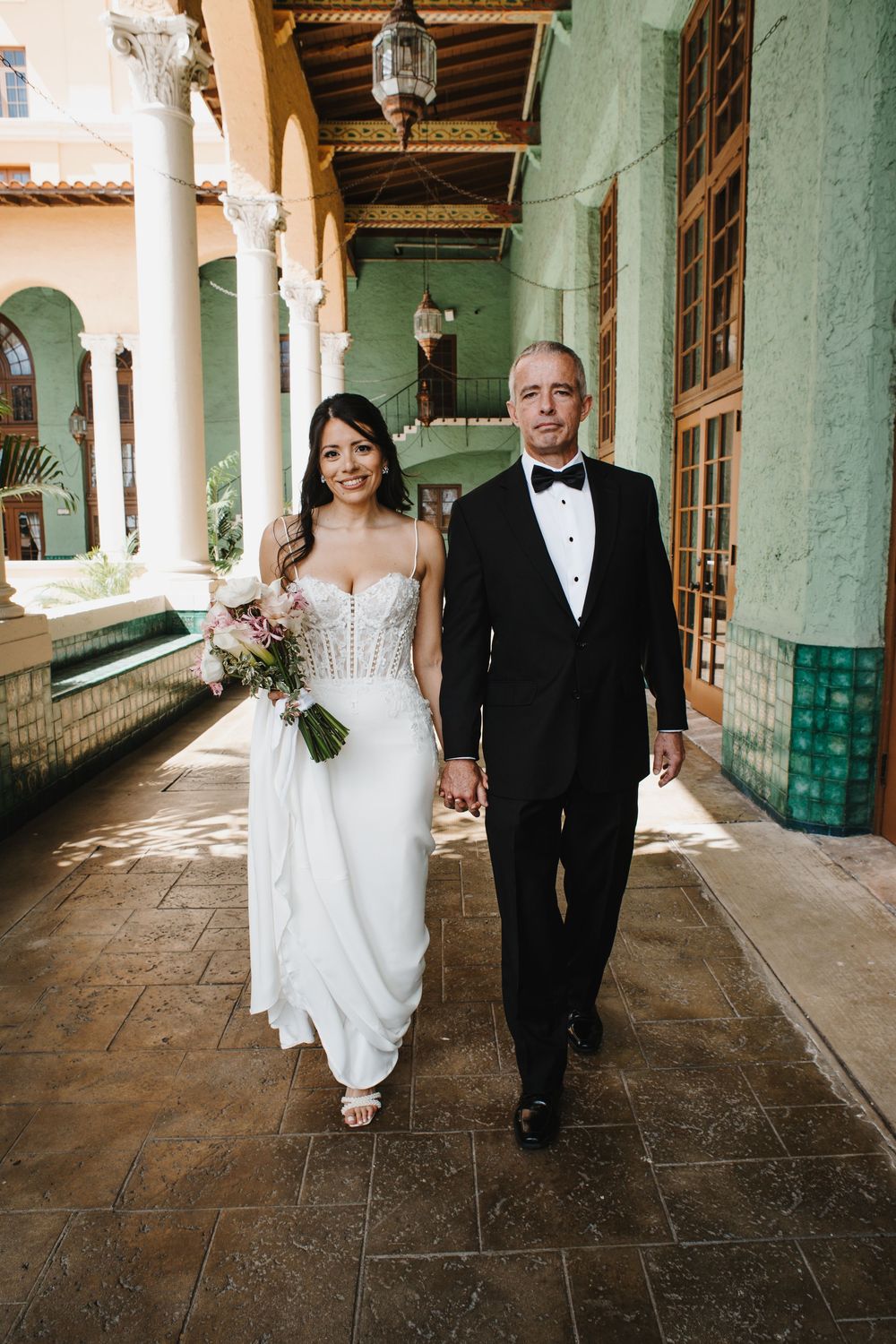 Bride holding bouquet and groom in tuxedo walk hand in hand through ornate hotel corridor.