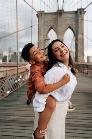 A joyful couple embraces and laughs together while standing on the iconic Brooklyn Bridge walkway in New York City.