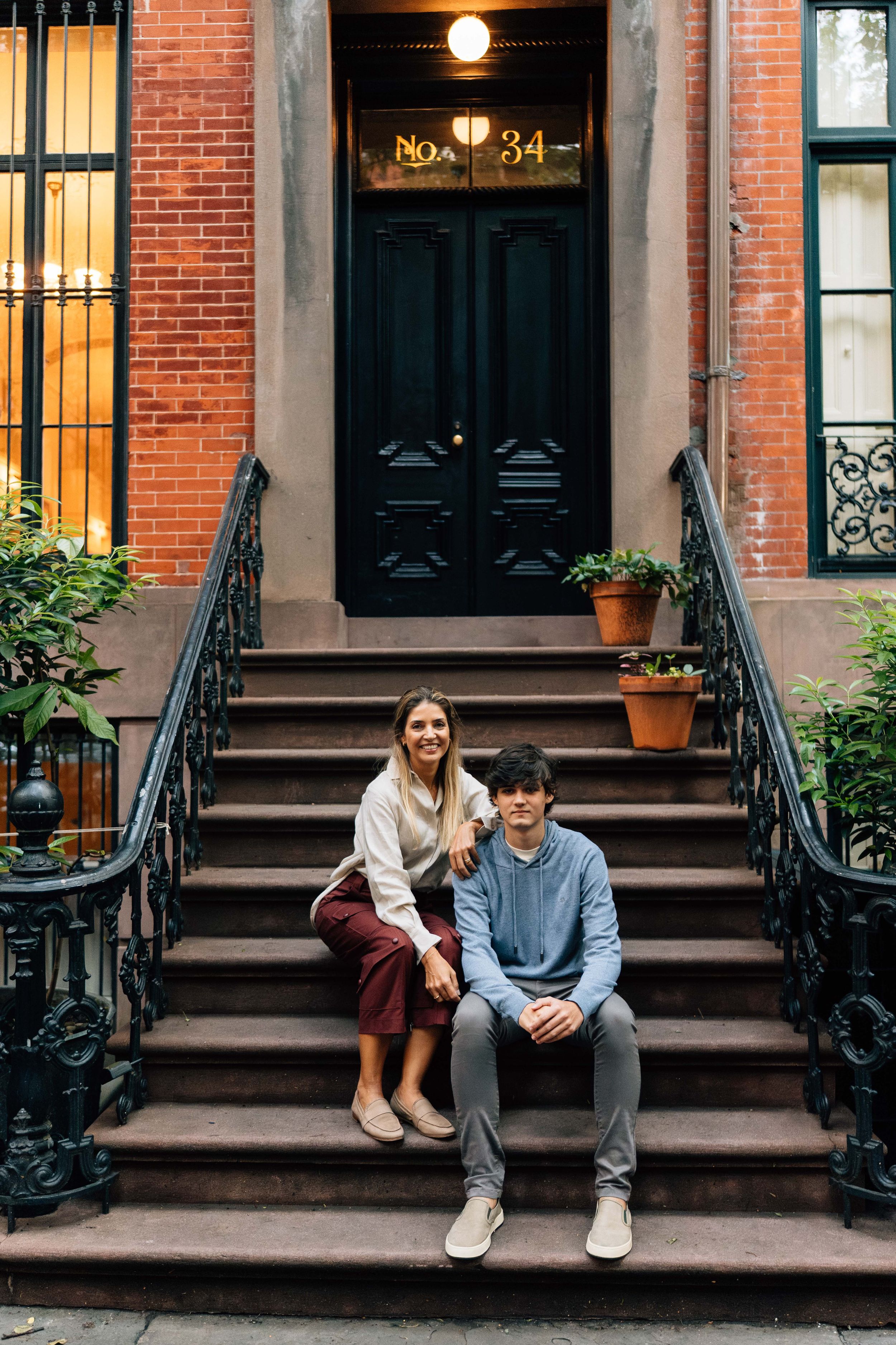 People sitting on brownstone steps in front of brick townhouse with black railings and ornate doors in New York City.