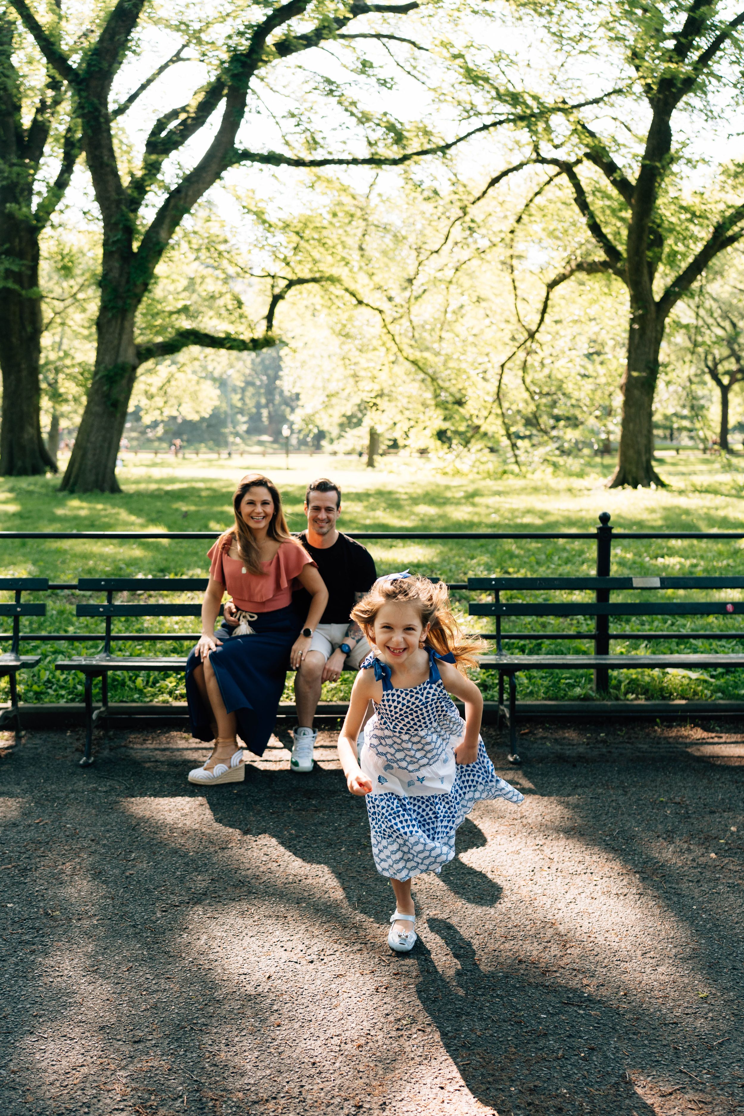 A young child runs joyfully on a sunlit path in Central Park while two people watch from a bench in the background.