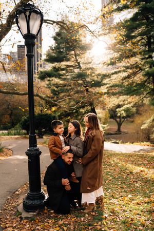 A group gathers around a vintage lamppost in a park during autumn with golden leaves scattered on the ground.