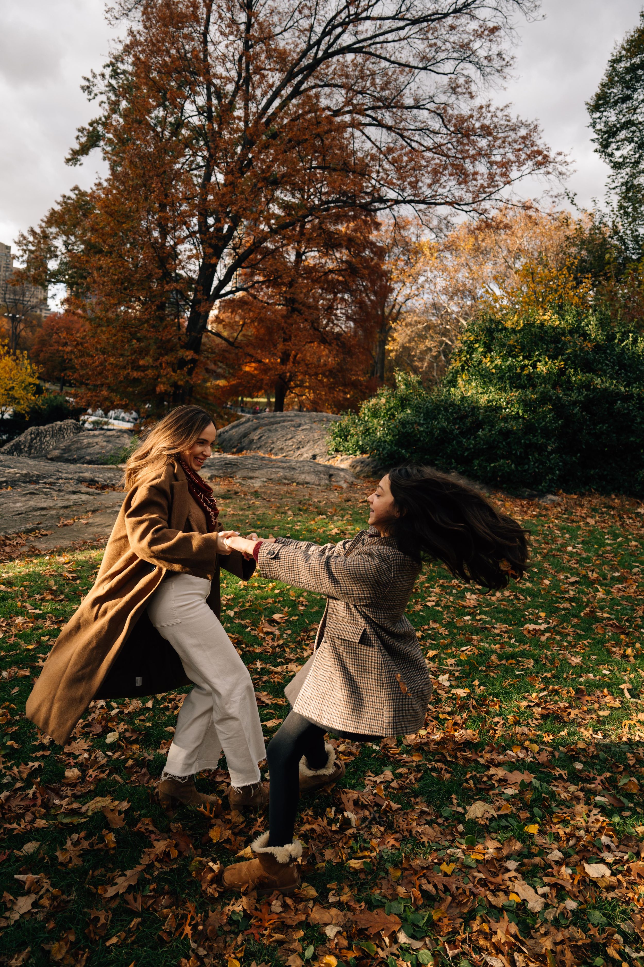 Two people in autumn coats dance and spin together on fallen leaves in a park with colorful fall foliage.