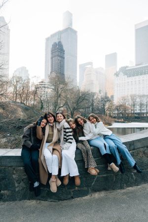 A group of friends sitting together on a wall in Central Park with the NYC skyline and skyscrapers visible in the background.